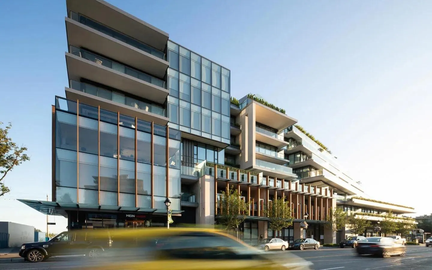Modern glass condo building with balconies and street-level retail. Cars drive by on the street in front.