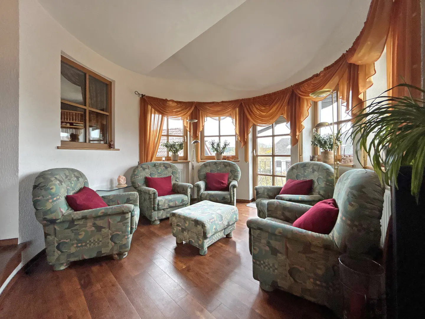 A curved living room features five armchairs with red pillows, a matching ottoman, and orange curtains.