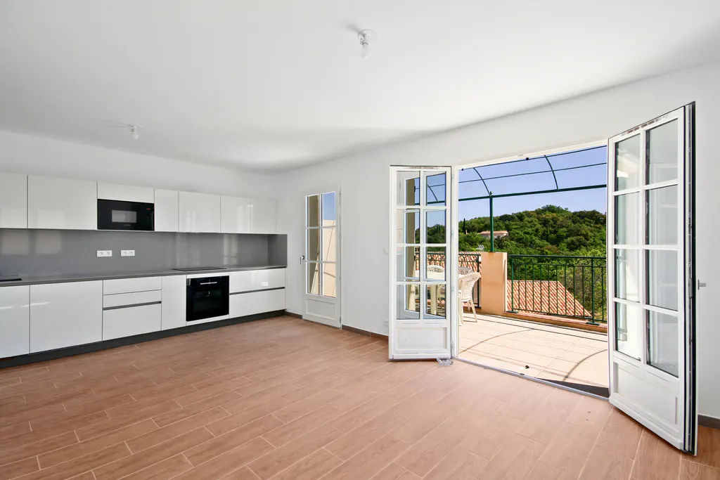 Bright, modern kitchen with white cabinets, wood floors, and open French doors leading to a balcony with green trees in the background.