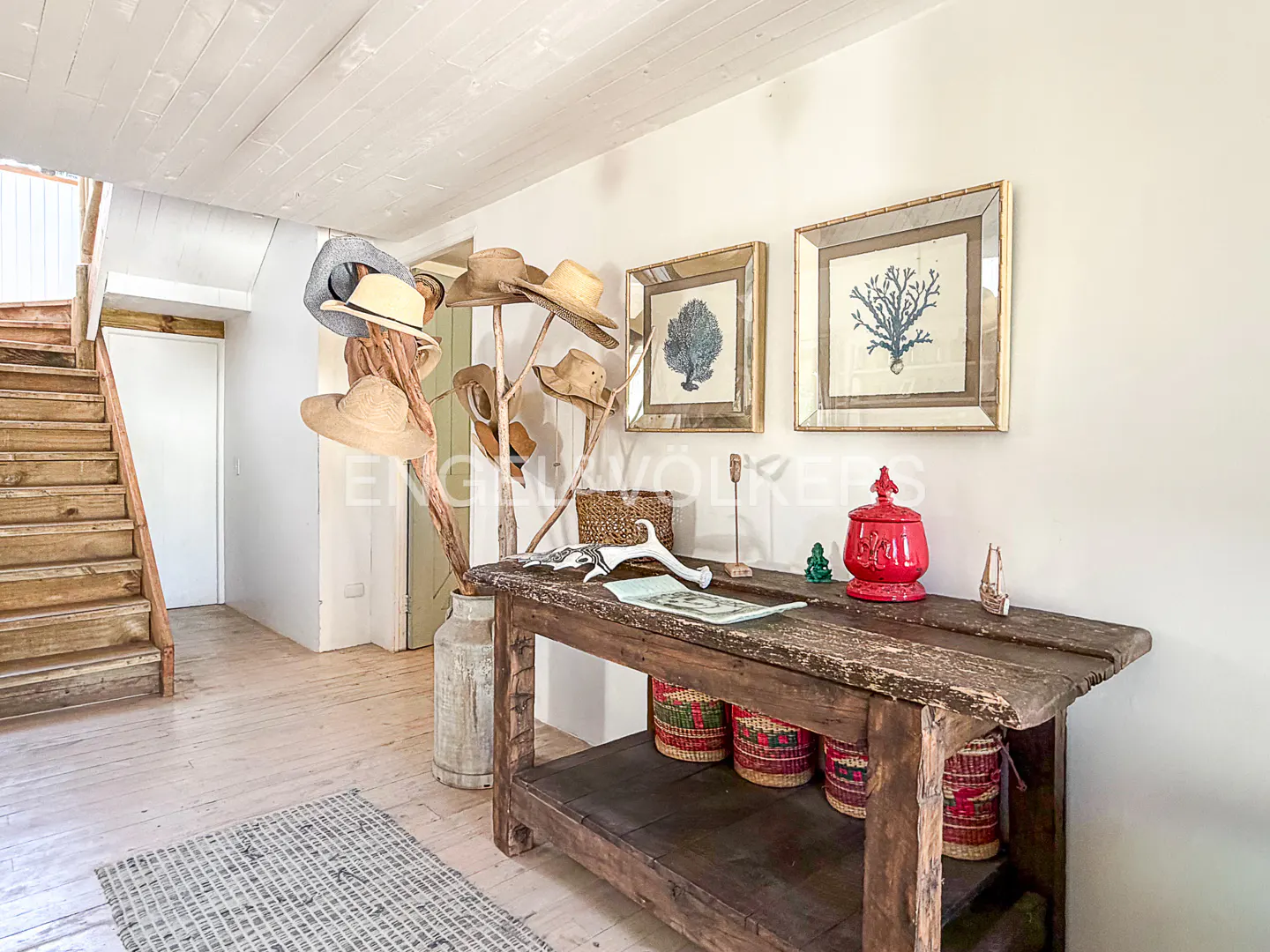 Rustic entryway with a wooden console table, a hat rack holding various hats, and framed coral art on a white wall.