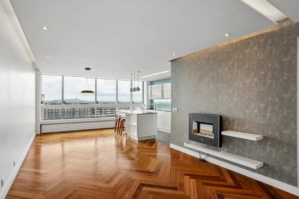 Bright, modern apartment with herringbone wood floors, a gray accent wall with a fireplace, and a kitchen island. Large windows overlook a cityscape.