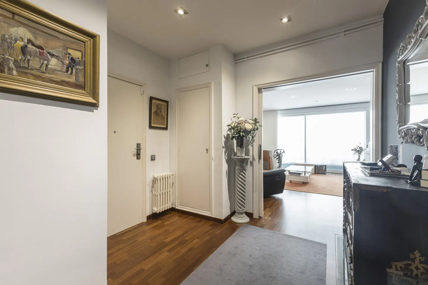 Entryway with hardwood floors, white walls, and a gray rug. A painting hangs on the left wall. A doorway leads to a bright living room.