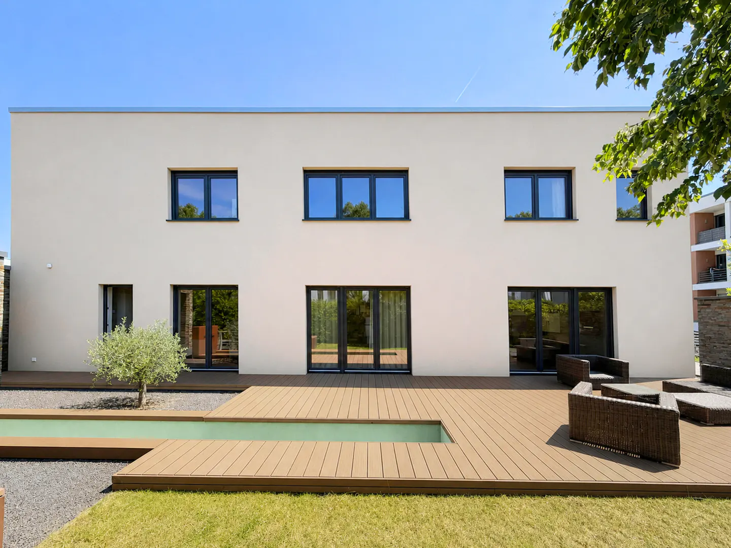 Modern home exterior with a beige facade, black-framed windows, and a wooden deck with outdoor seating. A small pool and green lawn add to the backyard's appeal.