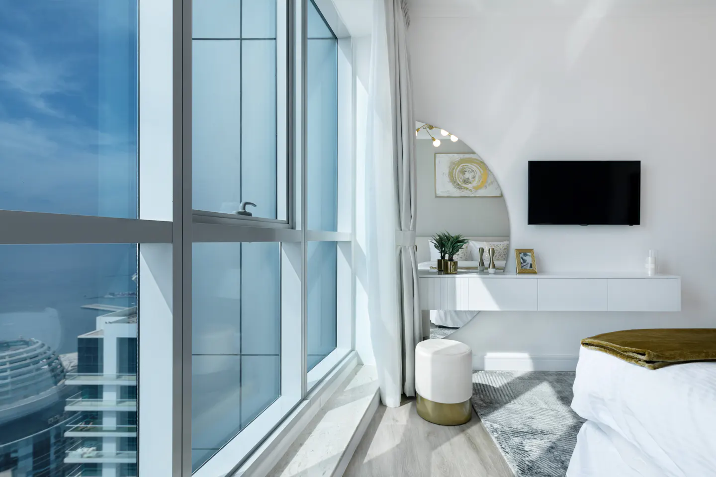 Bedroom with a large window showing a city view, a white vanity with a mirror, and a TV mounted on the wall.