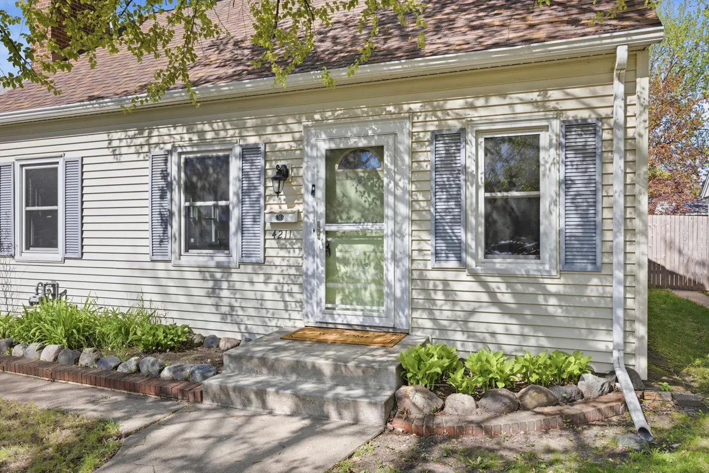 A one-story house with light yellow siding, gray shutters, and a brown roof. A green screen door leads to the front entrance.