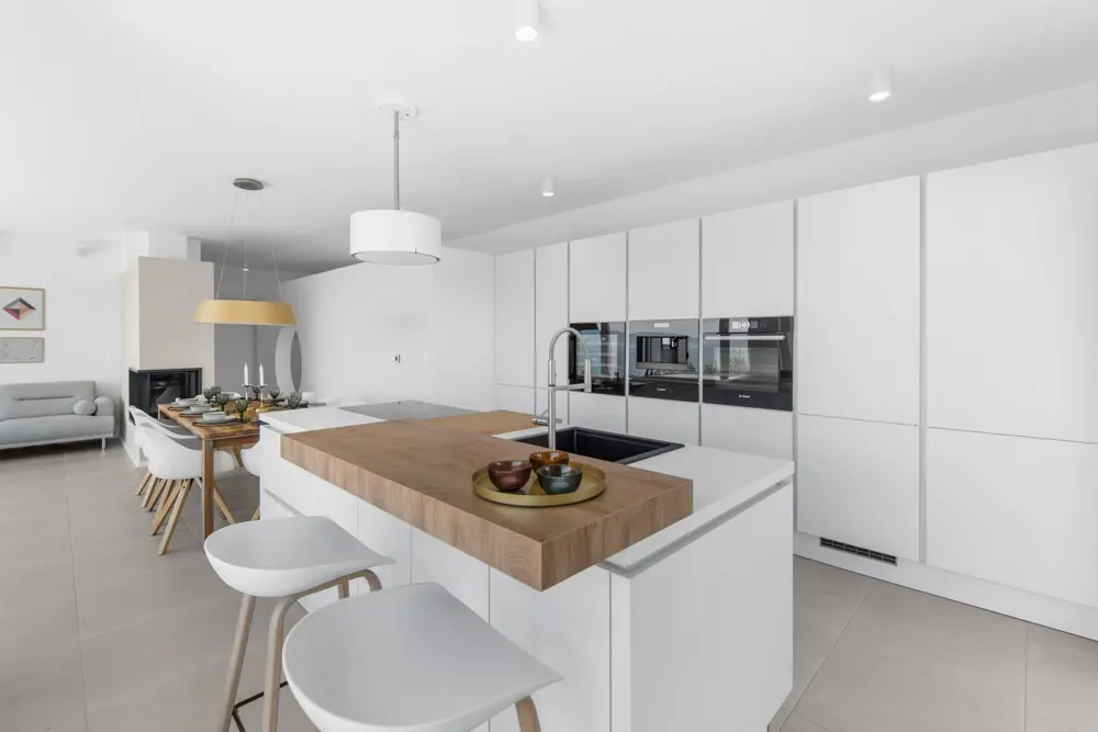 Bright, modern kitchen with white cabinets, wood island counter, and stools. Dining area and fireplace visible in the background.