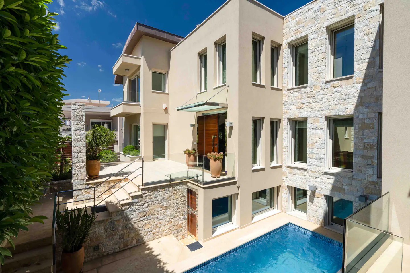Exterior view of a modern, multi-story home with a blue swimming pool. The house has beige stucco and stone accents, with many windows.
