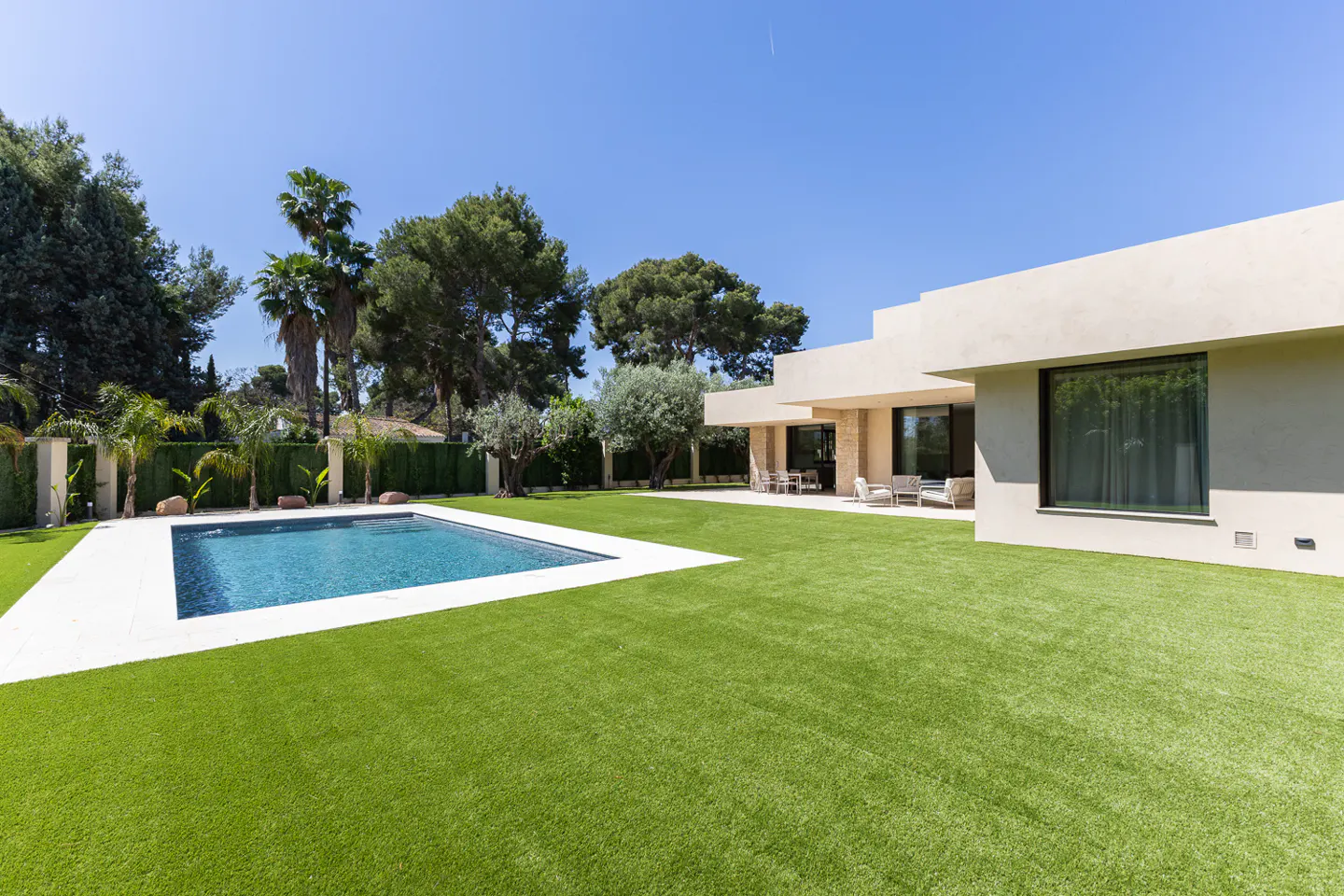 A modern home with a pool and green lawn under a blue sky. Palm trees and other greenery surround the property.