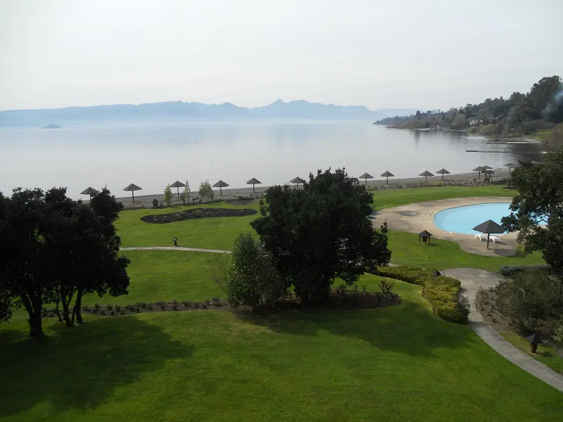 Scenic view of a lake with mountains in the distance, a beach with umbrellas, a pool, and green lawns.