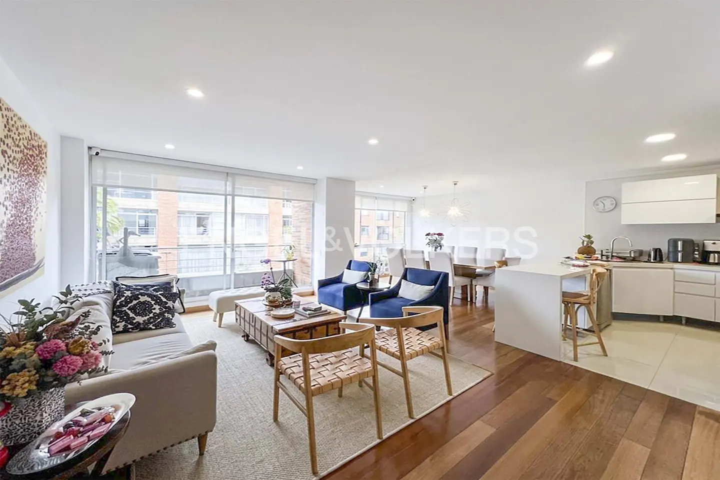 Open-concept living space with a beige sofa, blue chairs, wood floors, and a white kitchen. Large windows provide natural light.