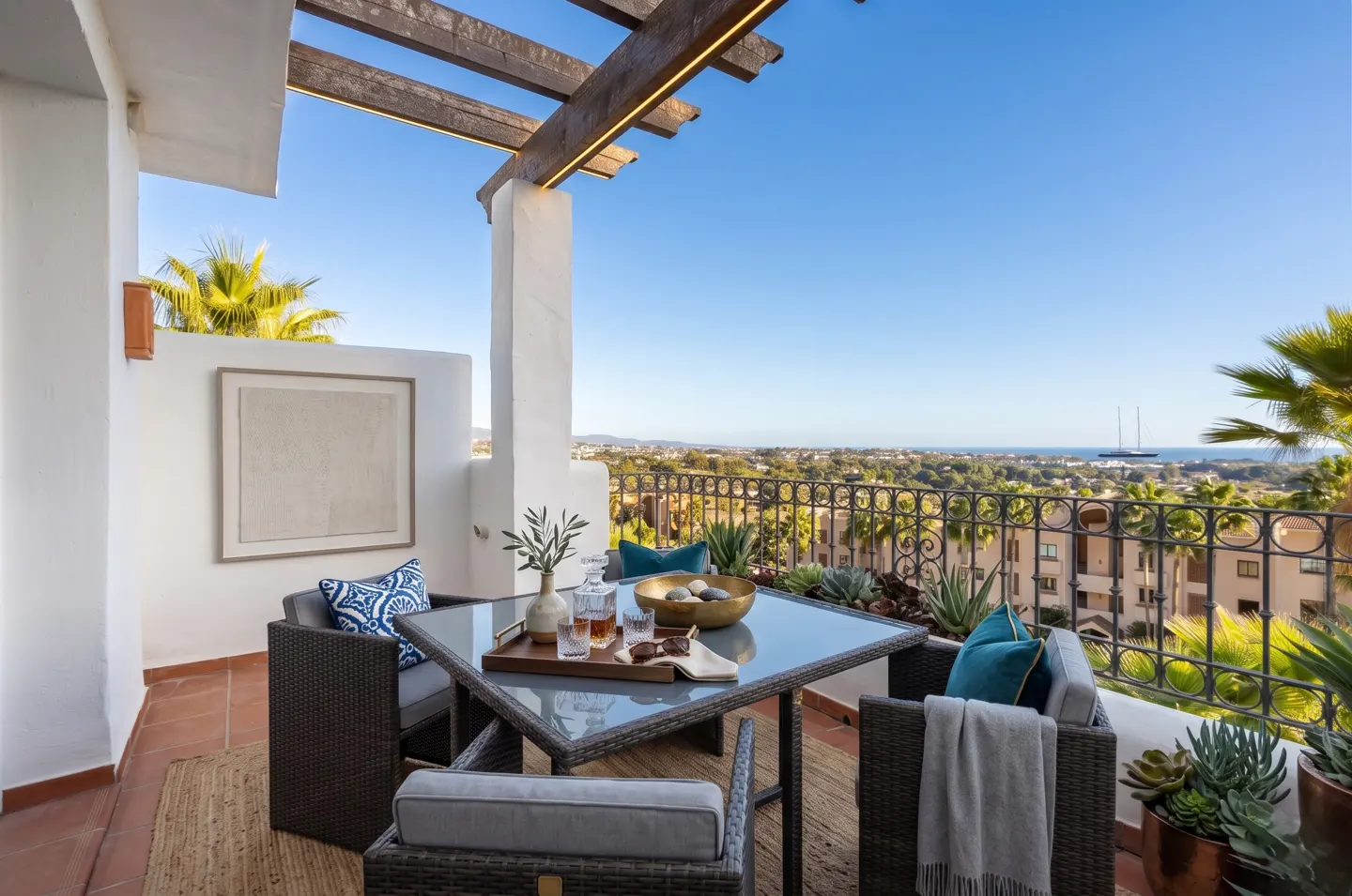 Outdoor patio with a glass table, wicker chairs, and blue pillows. A pergola covers the seating area, with a view of the city and ocean beyond the railing.