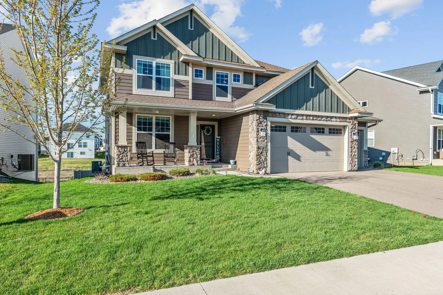 Two-story house with green and brown siding, stone accents, and a two-car garage. A green lawn and tree are in the front.