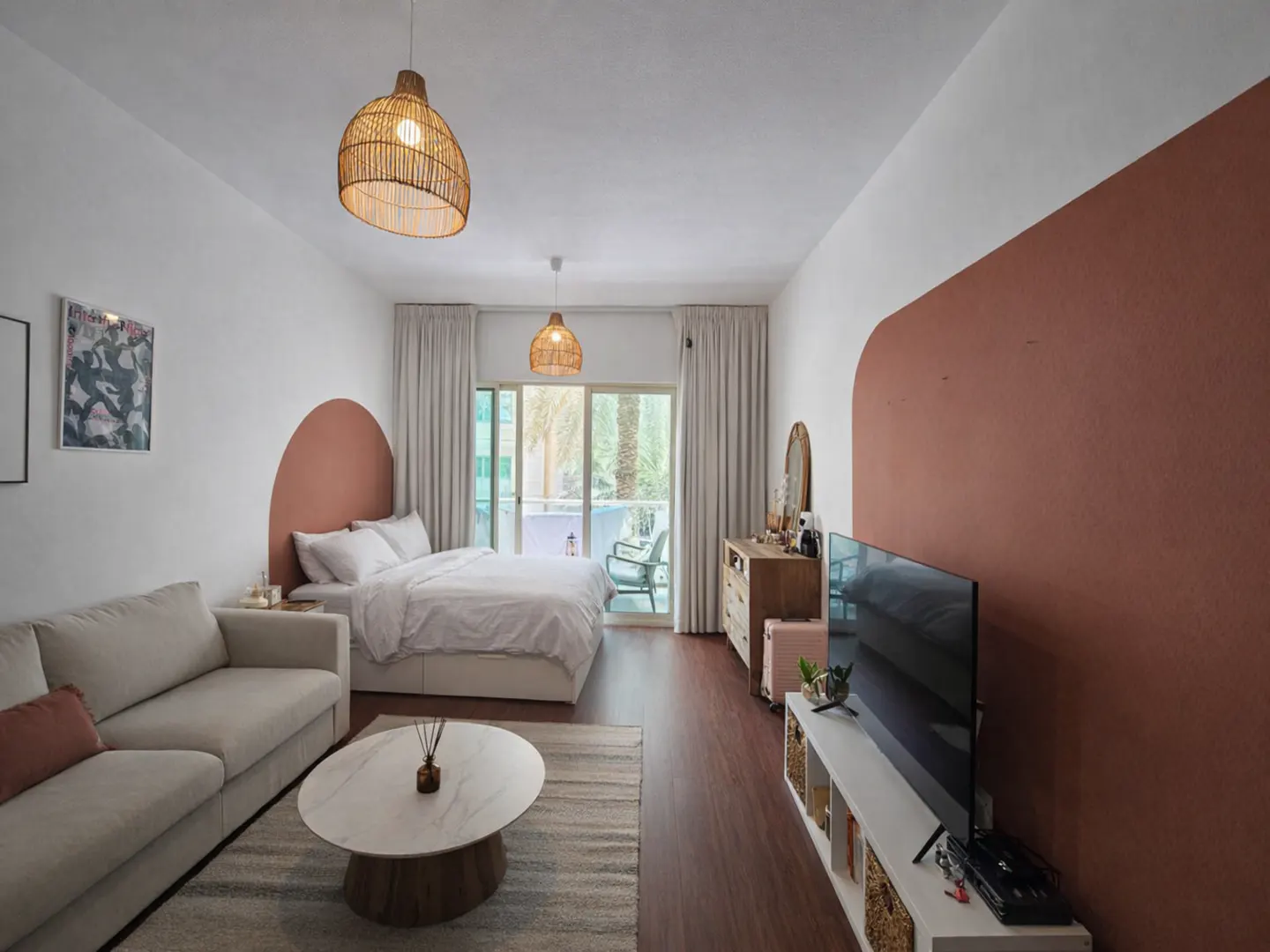 A bright studio apartment with a bed, sofa, TV, and balcony. The walls are white with terracotta arches. Rattan lamps hang from the ceiling.