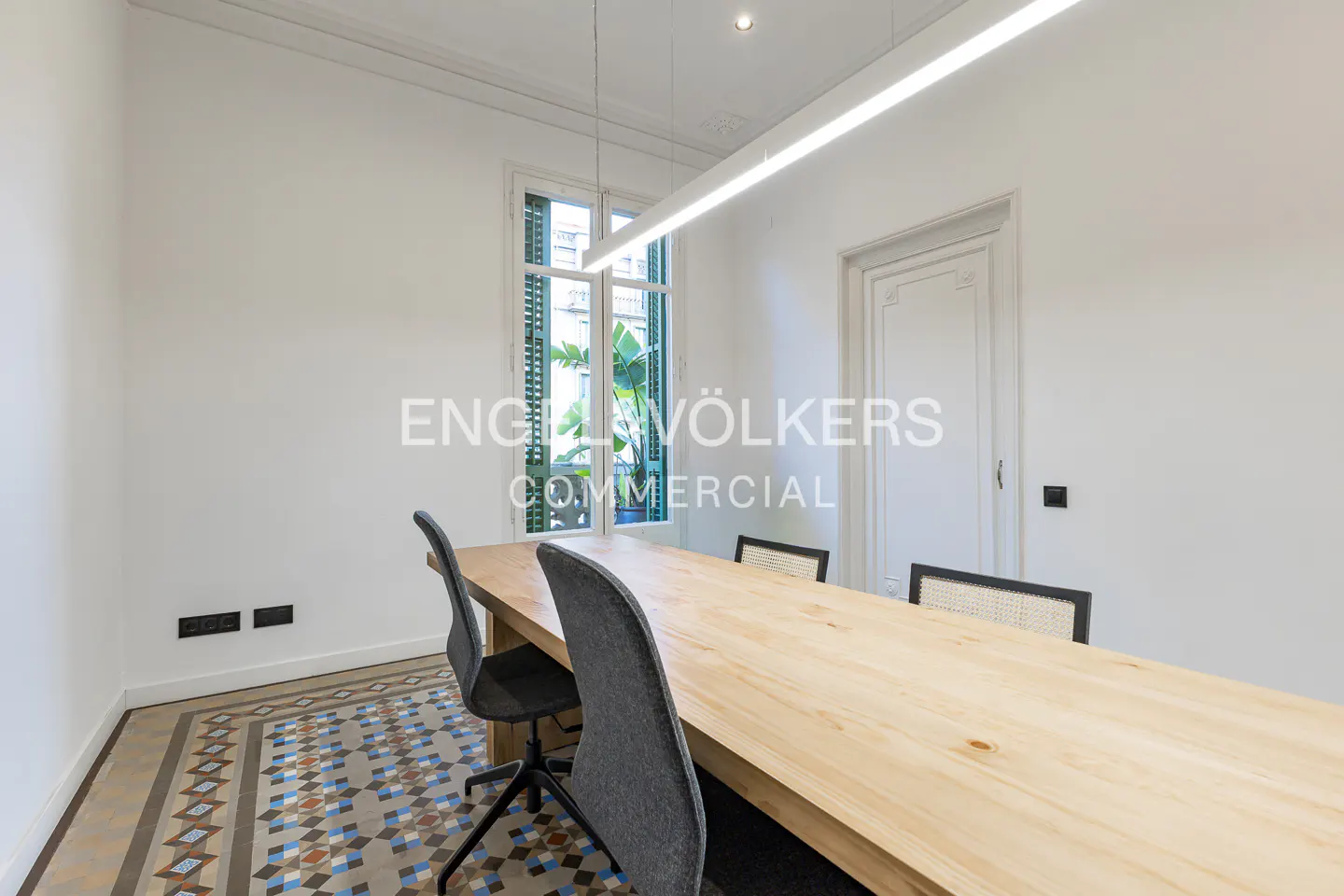 Bright office space with a long wooden table, gray chairs, and patterned tile floor. A window with green shutters is visible.