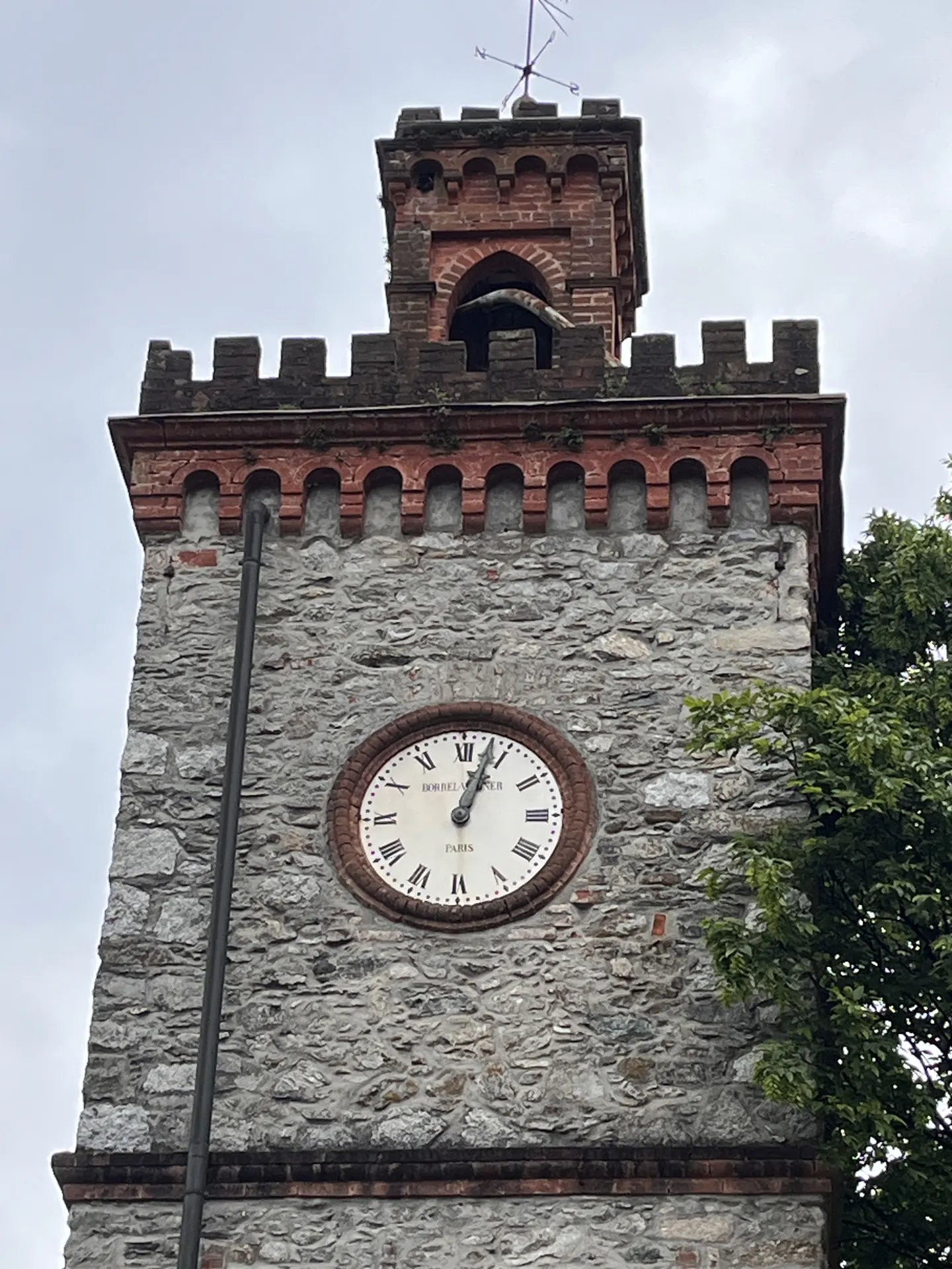 Close-up of a stone clock tower with a white clock face and Roman numerals, topped with a brick turret and weather vane.