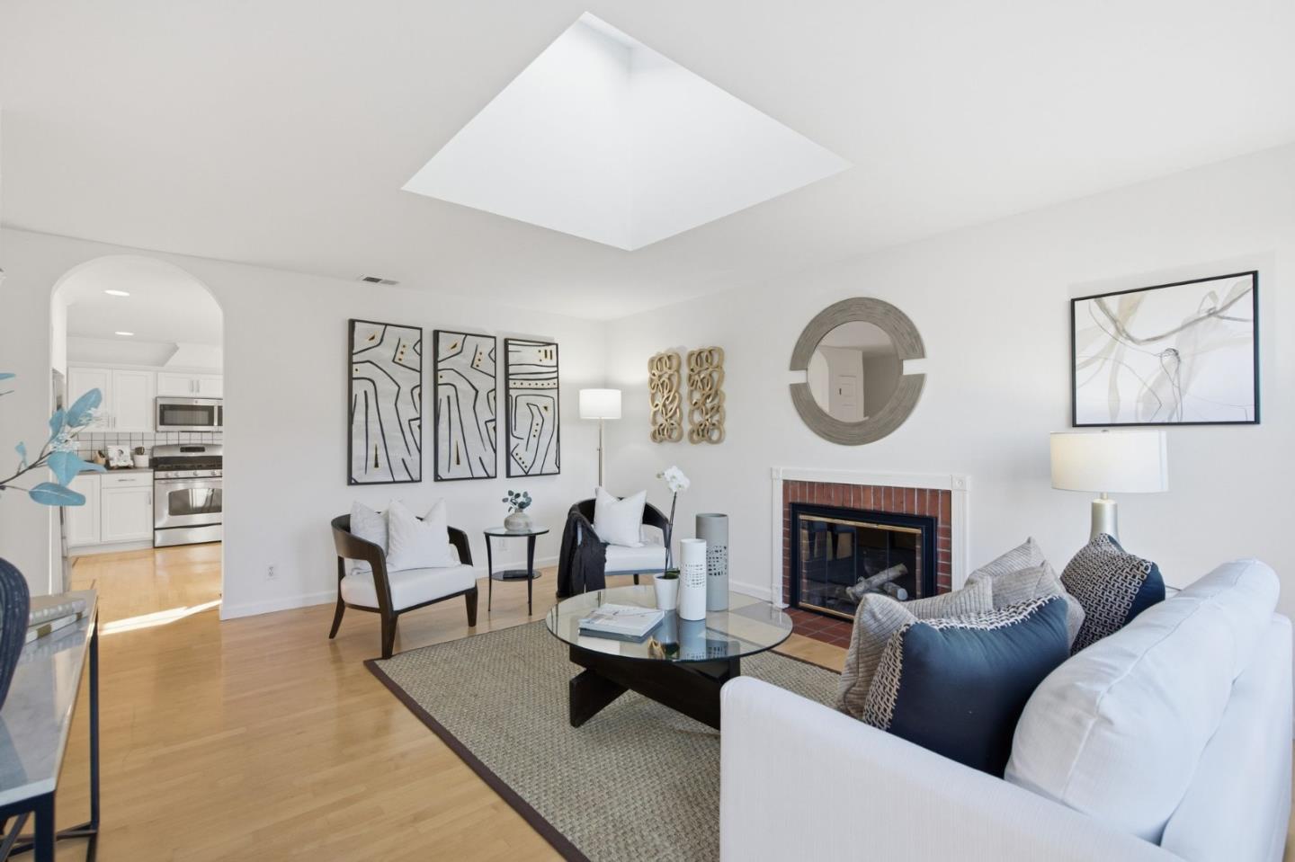Bright living room with white walls, wood floors, and a skylight. A white sofa, chairs, and a fireplace create a cozy atmosphere.