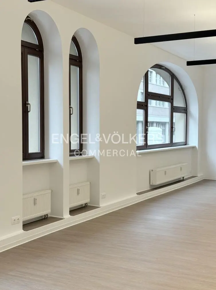 Bright, empty room with three arched windows, brown frames, white walls, and light wood floors. Radiators under windows.