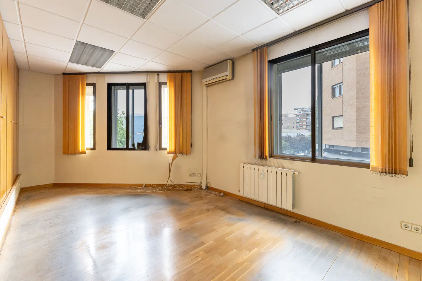 Empty office space with wood floors, beige walls, and black-framed windows with orange curtains. A radiator and air conditioner are visible.