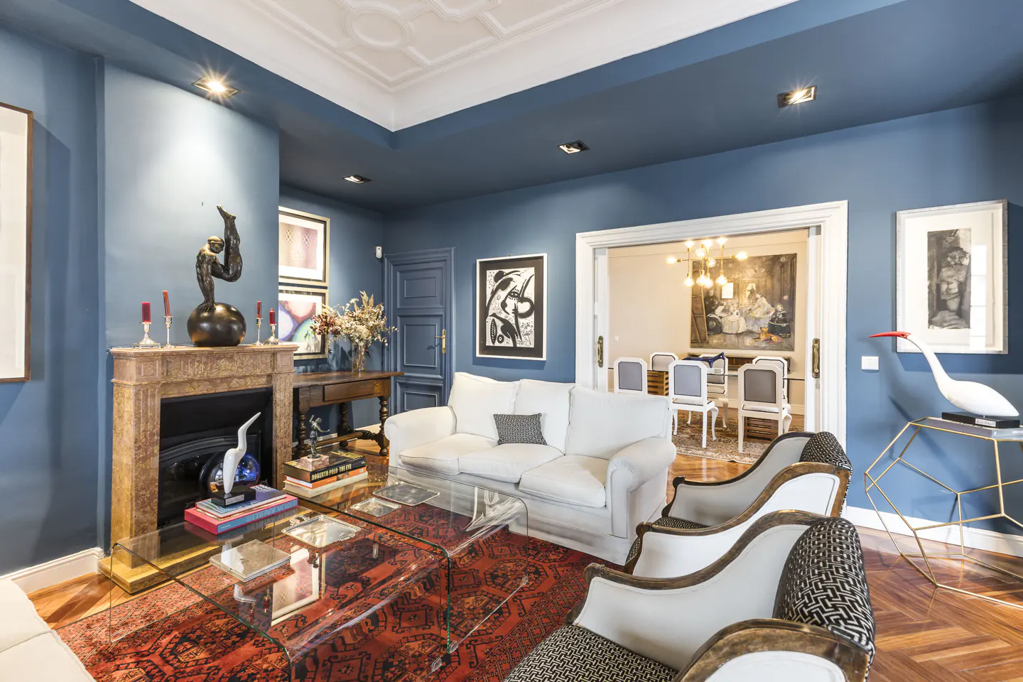 Living room with blue walls, white sofa, glass table on red rug, and dining room visible through doorway.