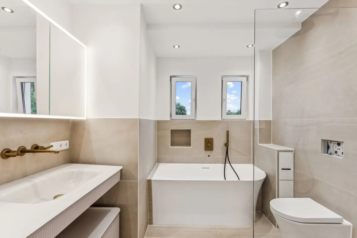Modern bathroom with beige tiles, a white soaking tub, wall-mounted brass faucet, and a sleek white vanity.