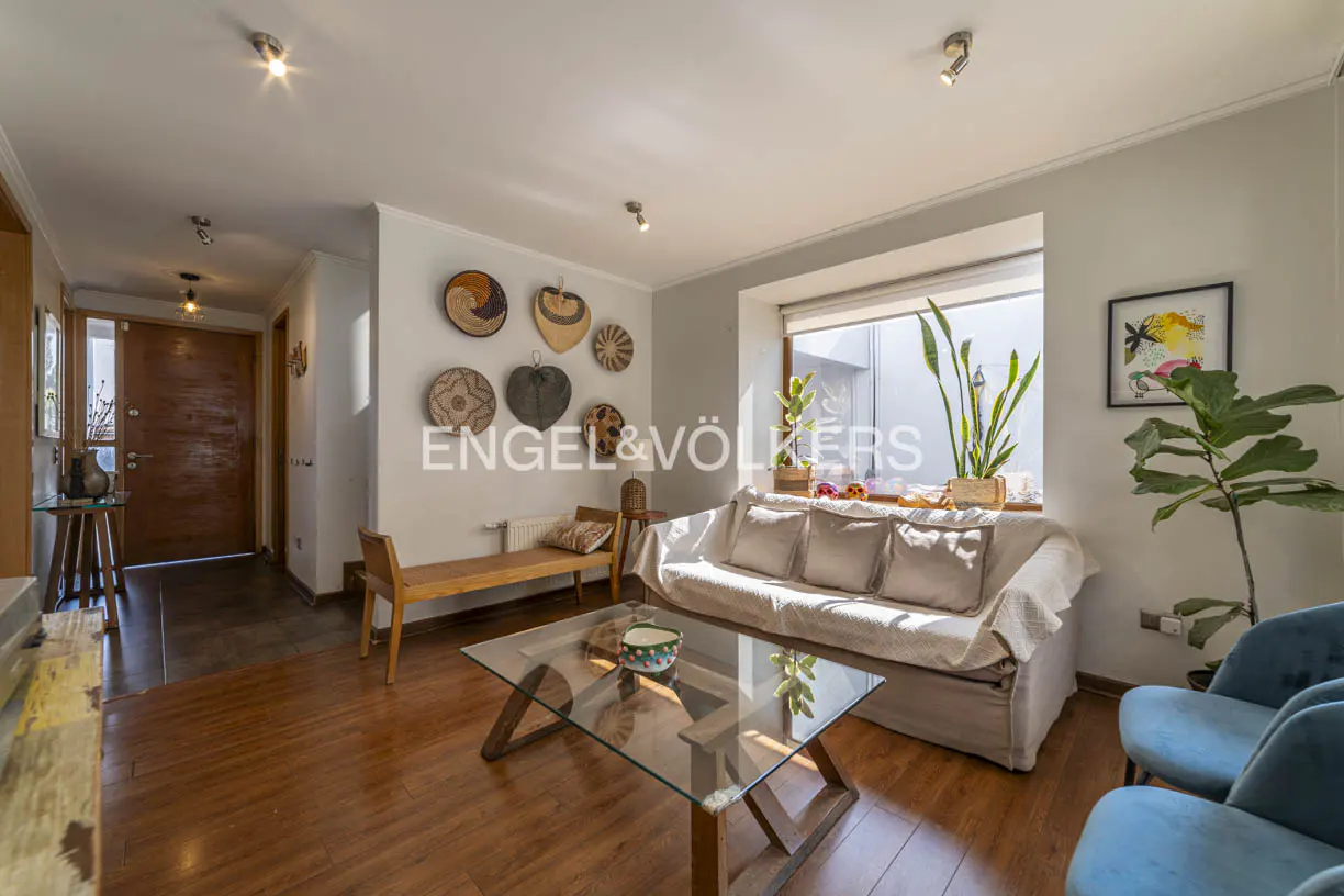 Bright living room with hardwood floors, a glass coffee table, a beige sofa, and blue armchairs. Woven baskets decorate the wall.