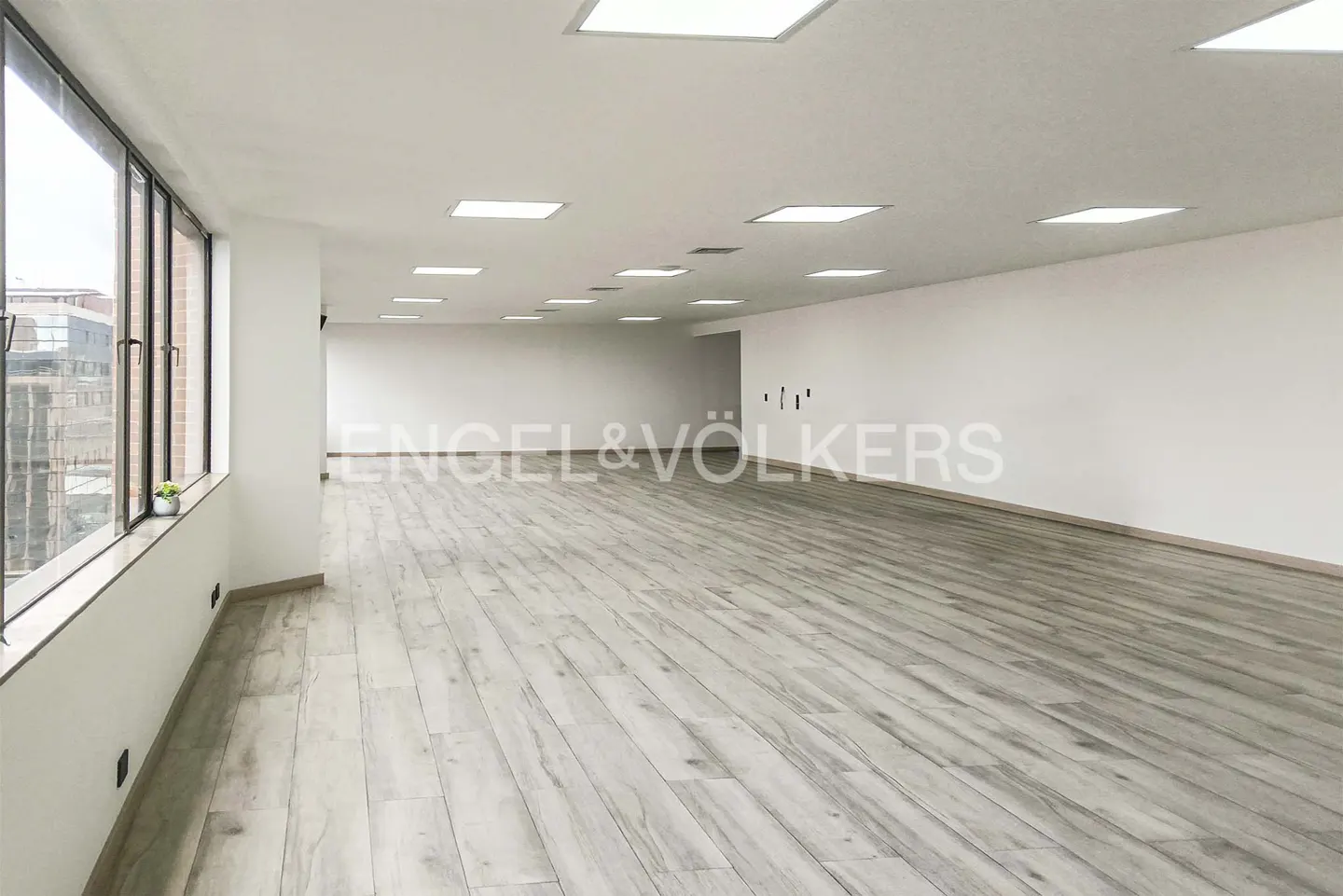 Bright, empty office space with wood-look floors, white walls, and large windows. Engel & Völkers logo on the wall.