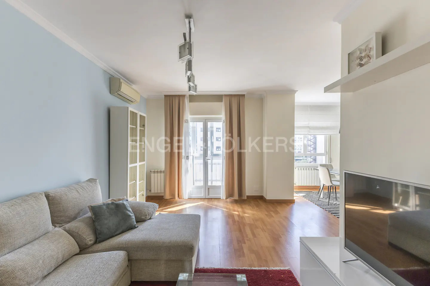A bright living room with a beige sectional sofa, hardwood floors, and a balcony with sheer curtains.