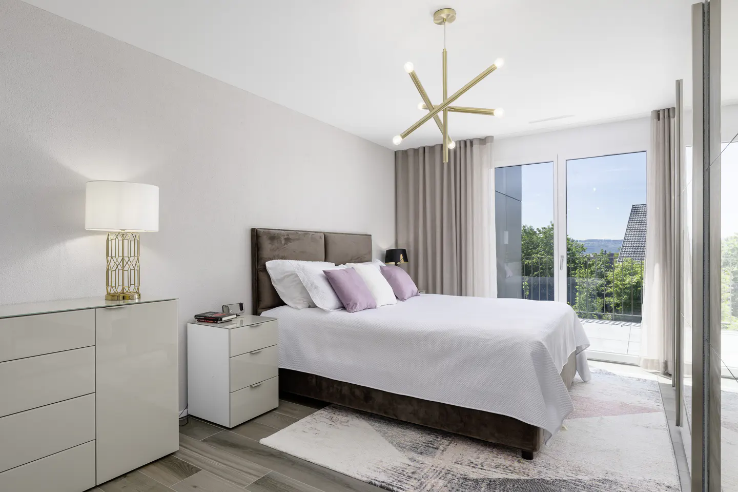 Modern bedroom with a brown upholstered bed, white bedding, purple pillows, gold chandelier, and a balcony with a view.