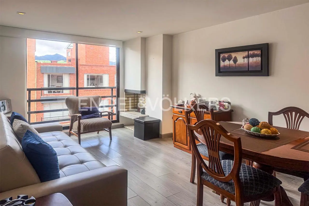 Living room with a beige sofa, striped armchair, wooden dining set, fireplace, and large windows overlooking brick buildings.