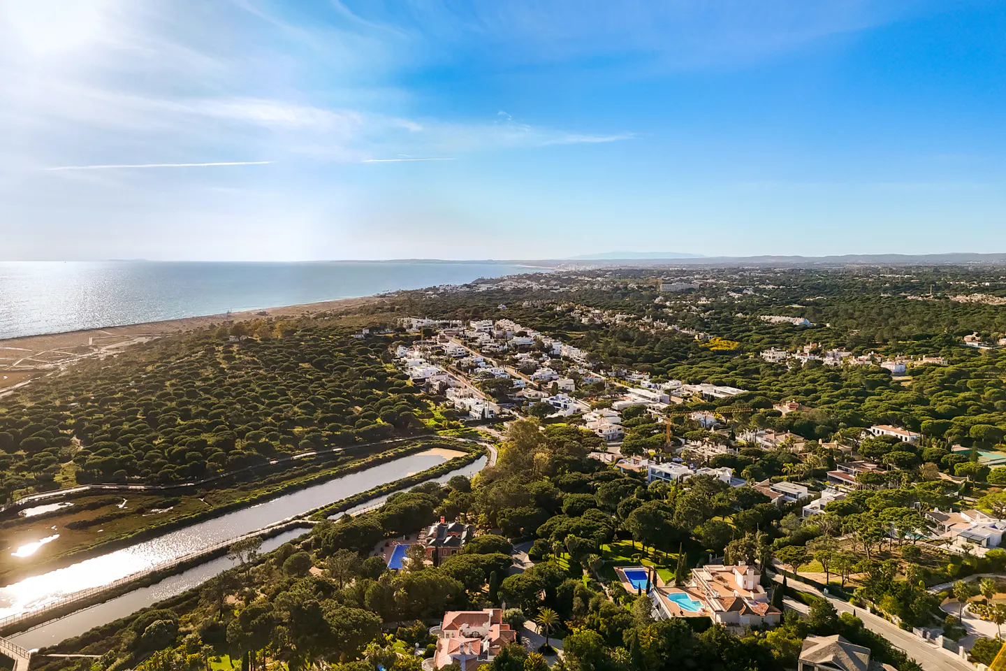 Aerial view of a coastal community with white houses nestled among green trees, near a beach and blue ocean under a bright sky.