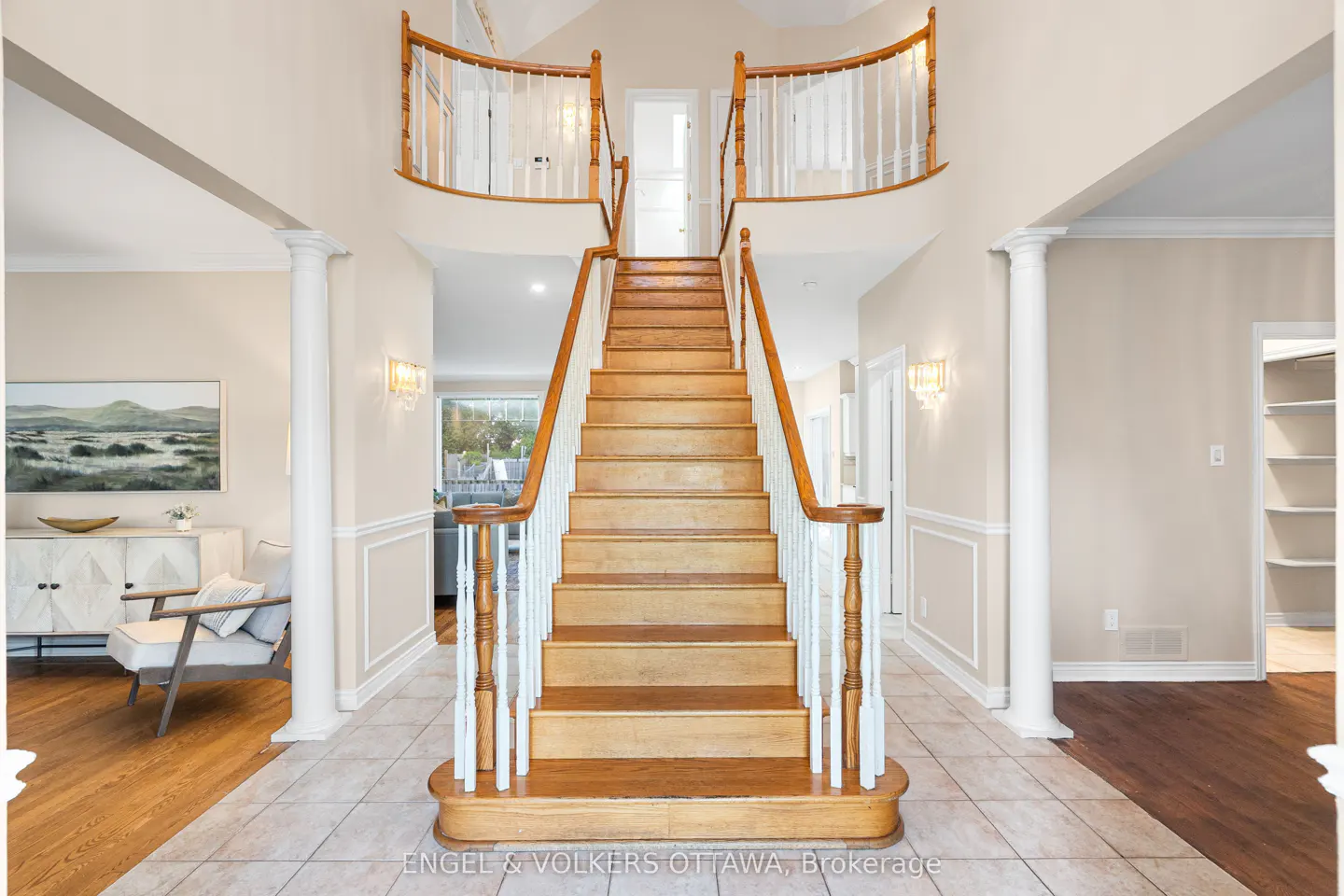 A bright foyer with a wooden staircase leading to a second-floor balcony with white railings. Cream walls, tile floor, and white pillars add to the spacious feel.