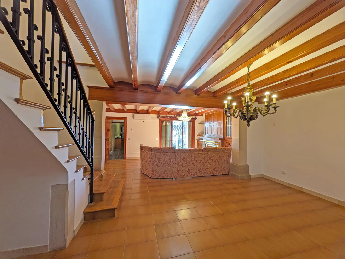 A spacious living room with terracotta tile floors, exposed wood beams, and a staircase with a black iron railing.