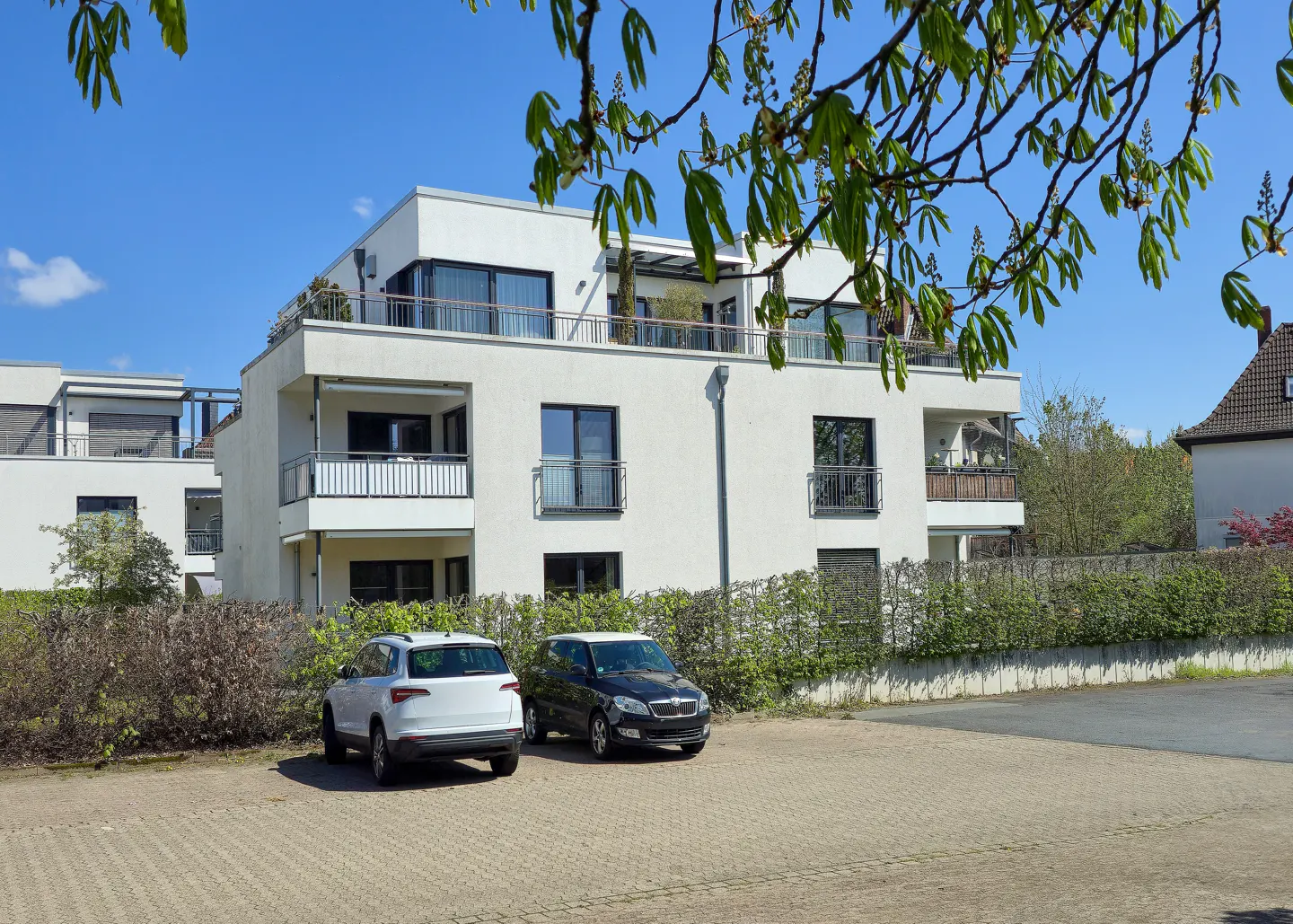 Modern white apartment building with balconies, parked cars, and green hedges under a blue sky.