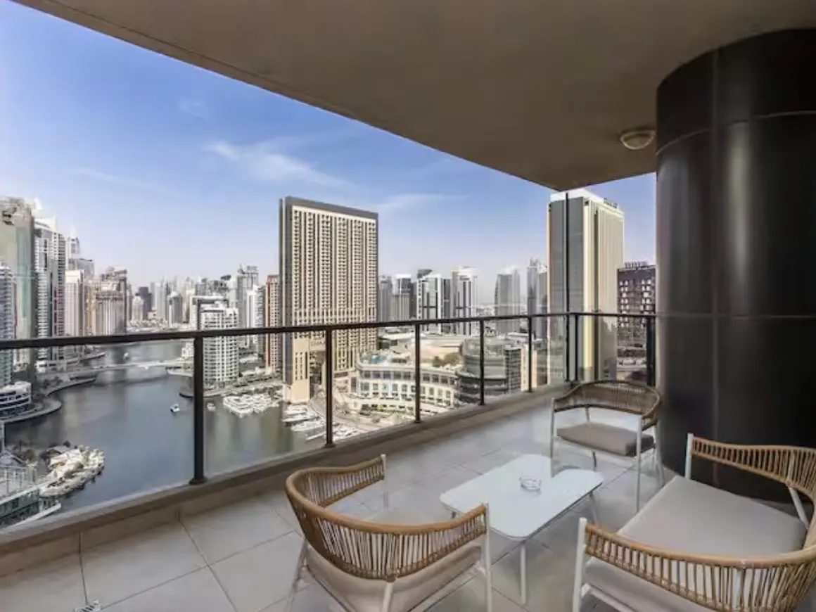 Balcony with wicker chairs and a white table overlooking a city skyline and a body of water.