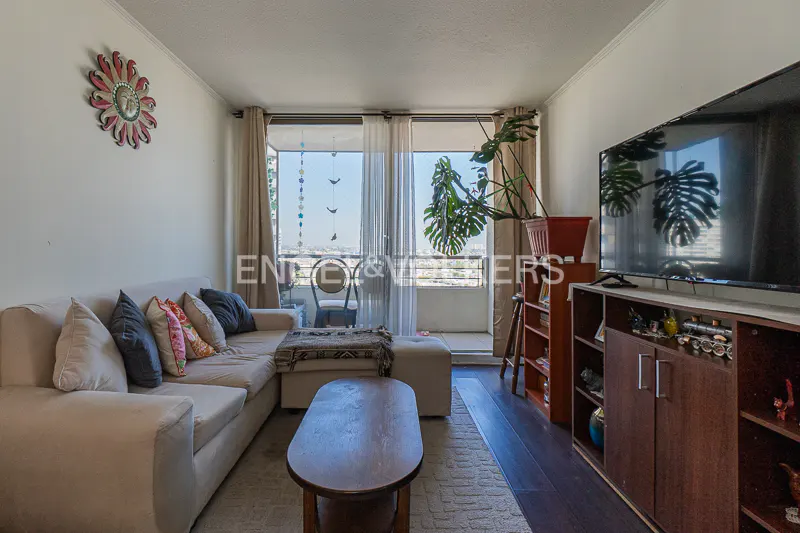 Living room with a beige sectional sofa, wood coffee table, TV, and balcony with city views. A sunburst wall decoration adds a pop of color.