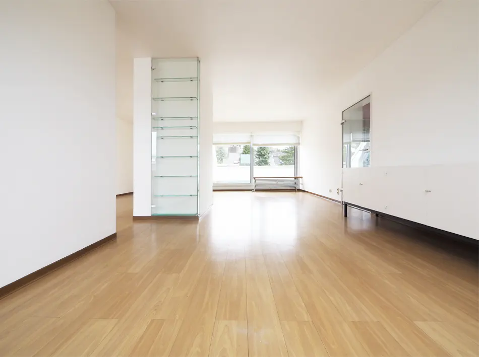 Bright, empty room with light wood floors and white walls. A glass shelf stands to the left, and a large window is in the background.
