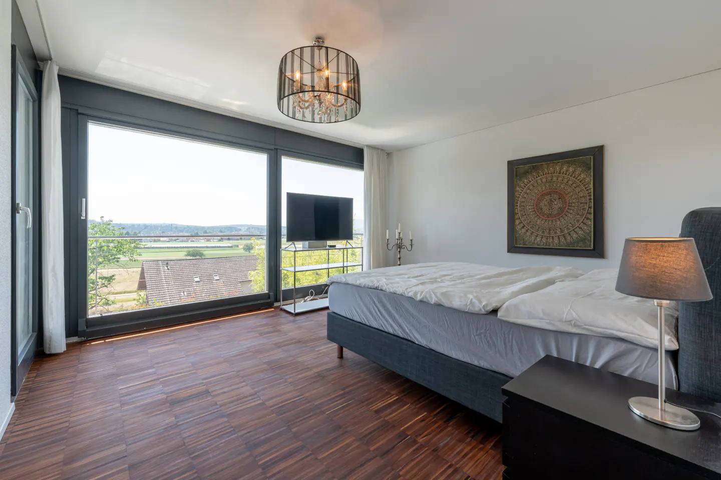 Bedroom with a large window view, wood floors, a gray bed, and a black nightstand with a lamp.