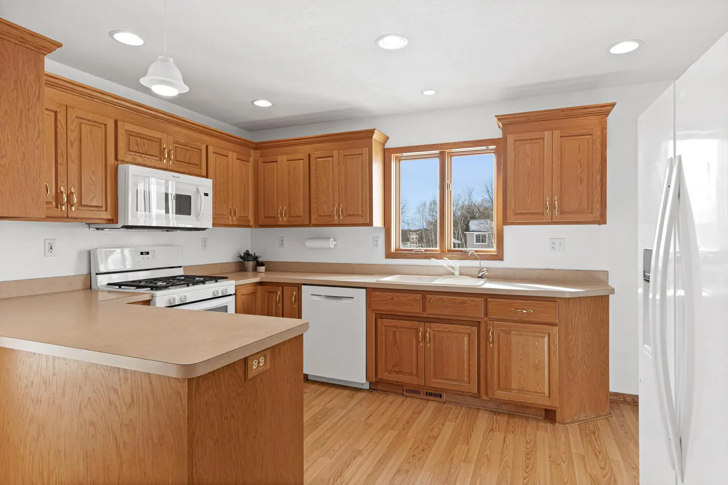 Bright kitchen with wood cabinets, light countertops, and wood floors. White appliances include a stove, microwave, dishwasher, and refrigerator.