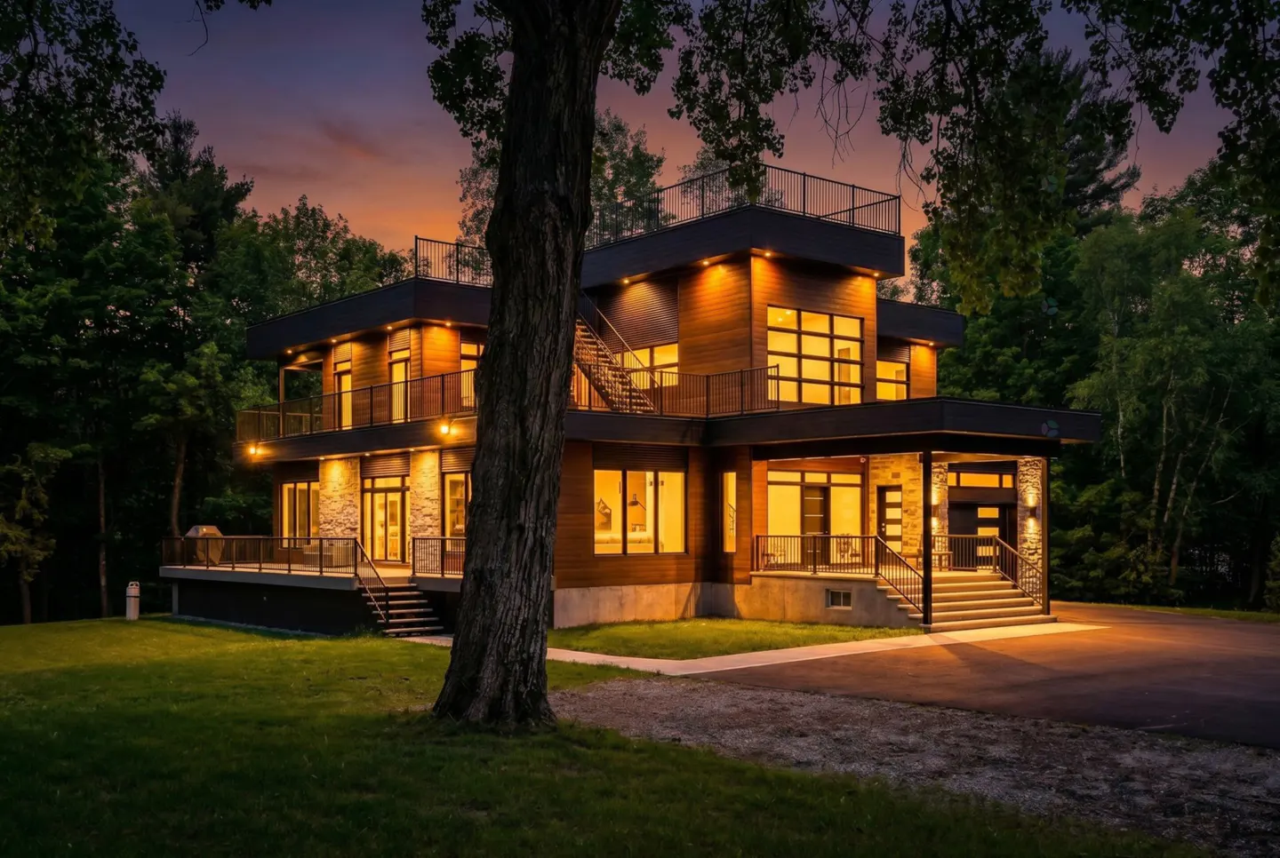 Modern multi-level home with wood siding and stone accents illuminated at dusk, surrounded by trees and a grassy lawn.