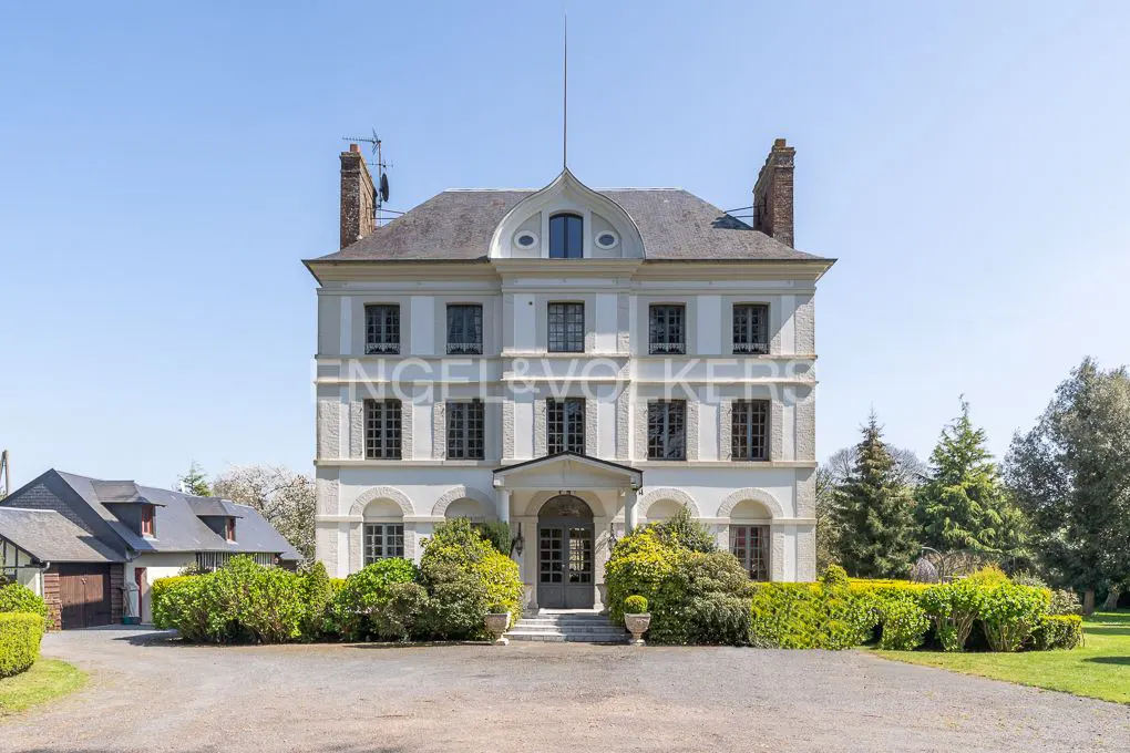 Exterior view of a three-story white mansion with a gray roof, dark windows, and green landscaping on a sunny day.