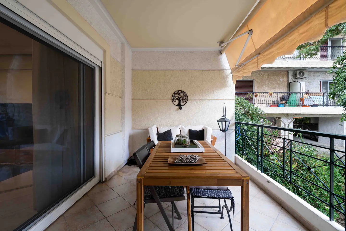 A balcony with a wooden table and chairs, a white sofa, and a black metal tree decoration on the wall. A black metal railing overlooks greenery.