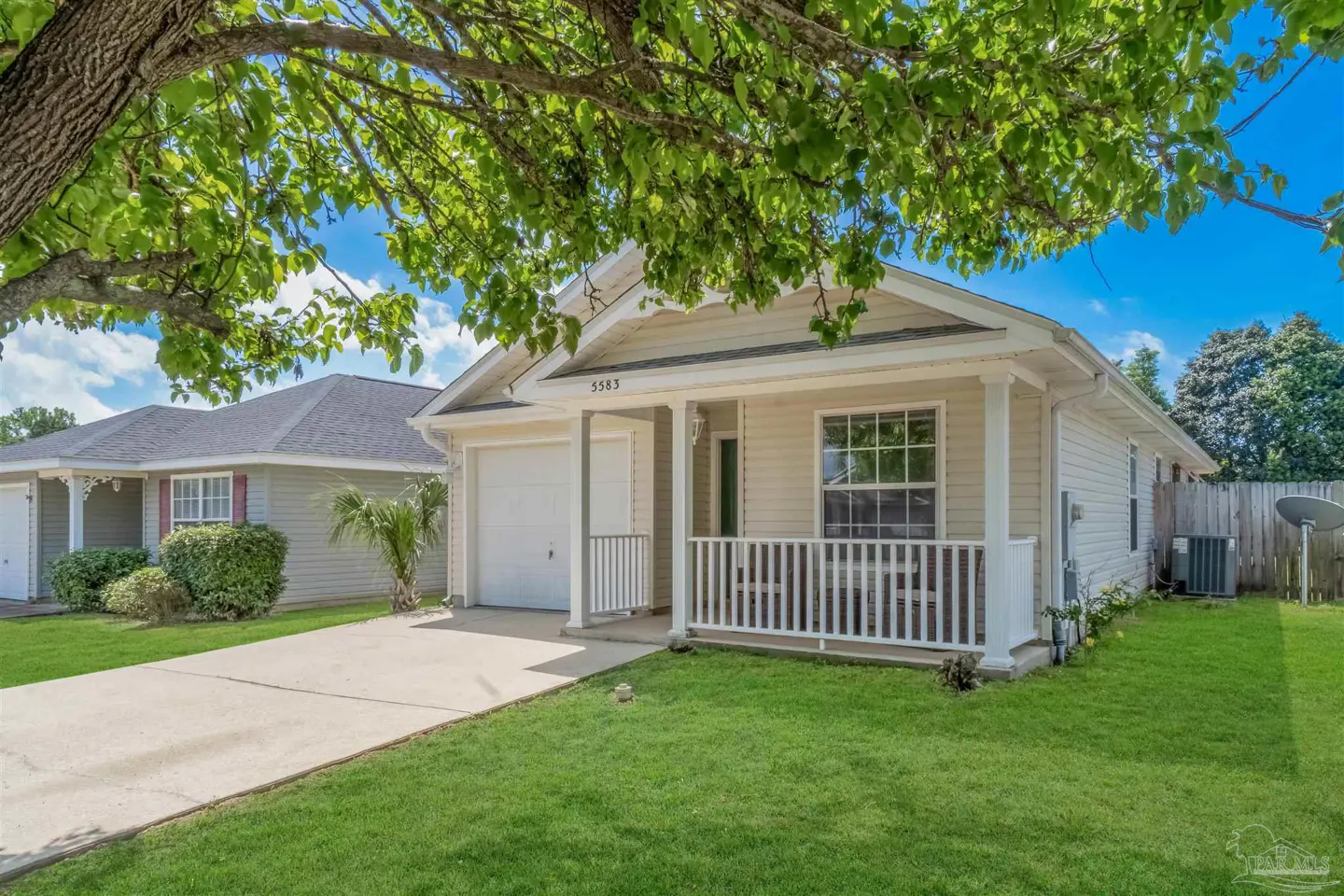 Beige single-story house with a white garage door and porch railing, green lawn, and tree branches overhead.