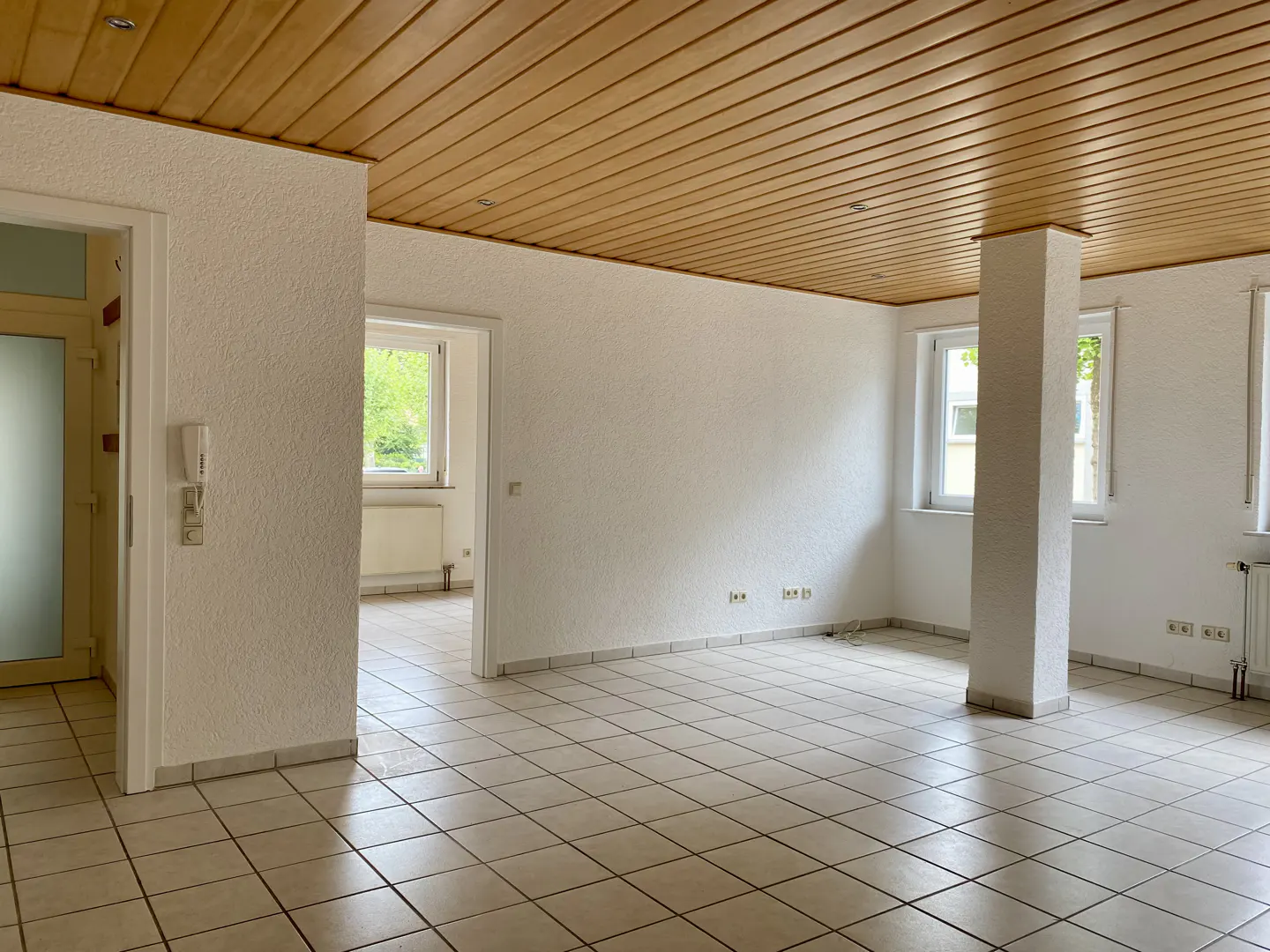 An empty room with white walls, a wood-paneled ceiling, and a tiled floor. There are windows and doorways visible.