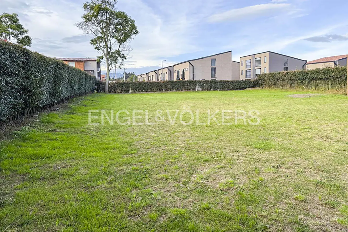 A grassy lot with a hedge border and modern houses in the background. The sky is partly cloudy.