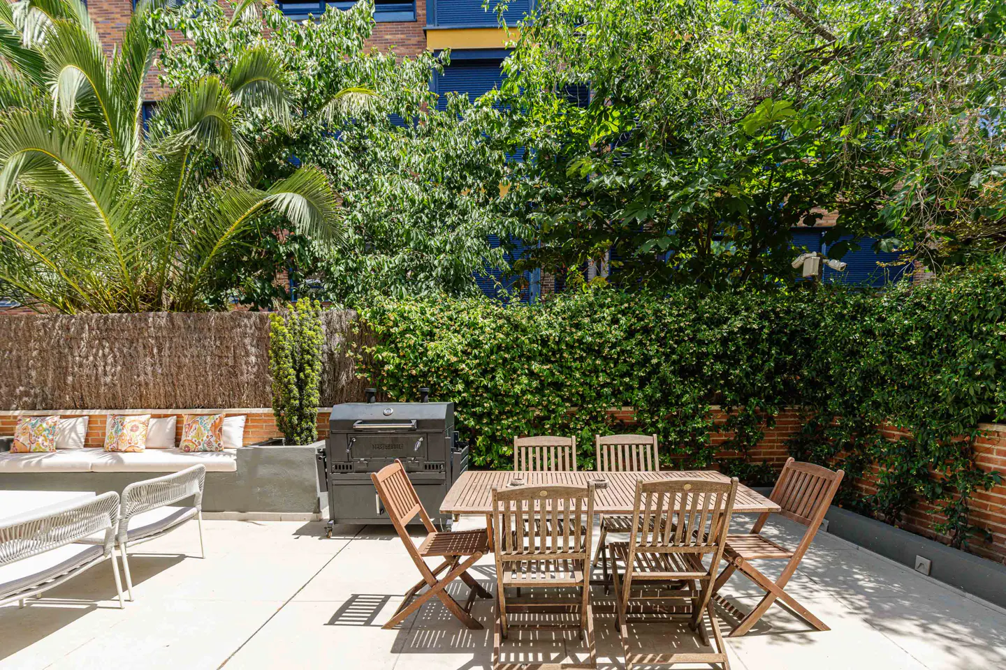 Sunny patio with a wooden dining set, a black grill, and a cushioned bench, surrounded by lush green trees and hedges.