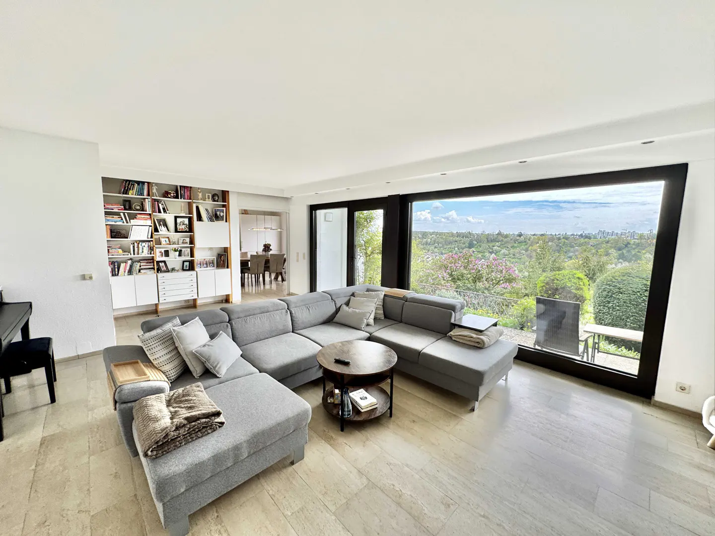 Bright living room with a gray sectional sofa, round coffee table, and a large window with a view of trees and sky. A bookshelf and piano are visible.