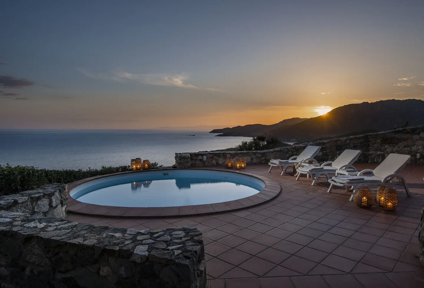 Sunset view of a round pool with lounge chairs, lanterns, and ocean backdrop.