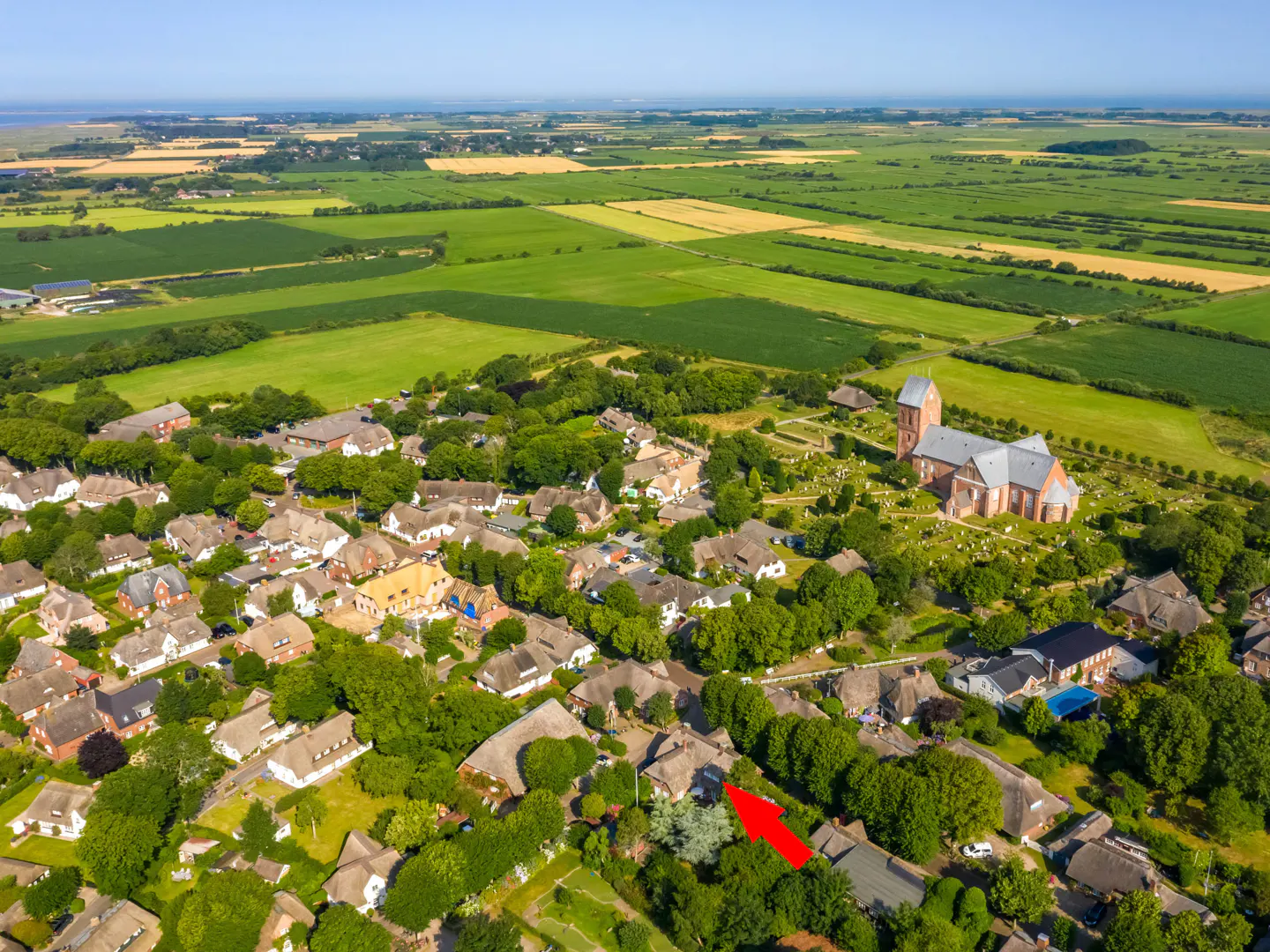 Aerial view of a village with thatched-roof houses, green trees, and a red brick church in the background. A red arrow points to a specific house.