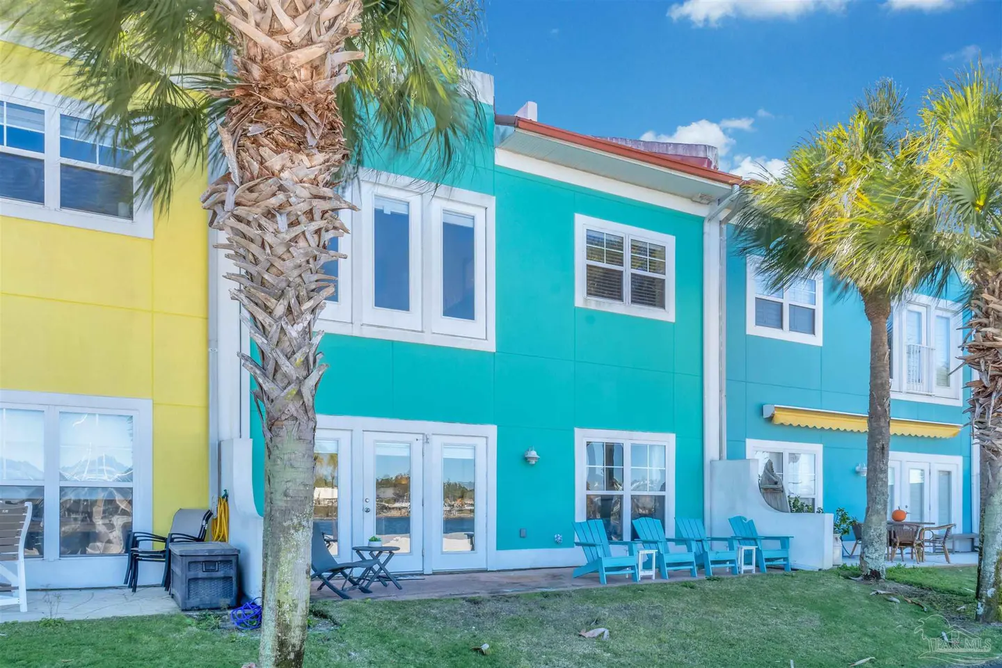 Exterior view of colorful townhouses with yellow and turquoise walls, white trim, and palm trees. Blue chairs sit on the lawn.