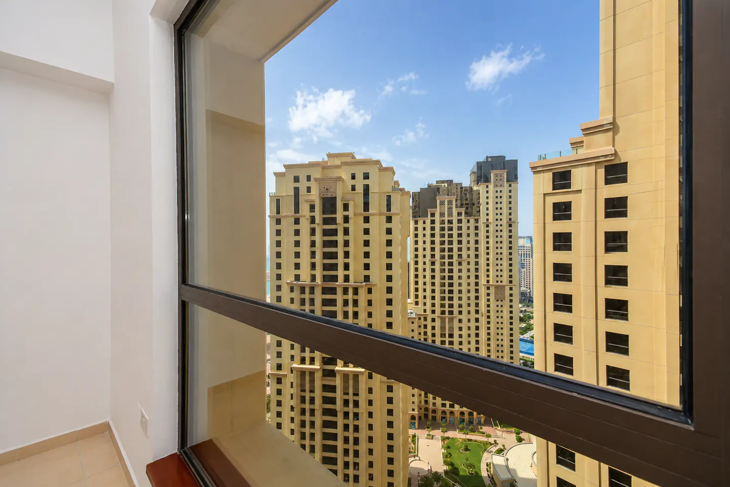 View from a window of tall, tan buildings under a blue sky with white clouds. The window frame is dark brown.